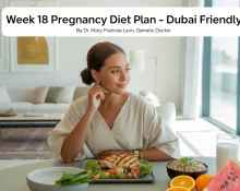 A health-conscious pregnant woman relaxes in a sunlit, modern Dubai apartment. She wears breathable cotton loungewear and sits at a dining table with a light meal: grilled chicken salad, a small bowl of sliced fruits, and a glass of coconut water. The apartment features minimalist décor with Arabic design elements, gold accents, and indoor plants. The setting is calm, fresh, and wellness-focused.
